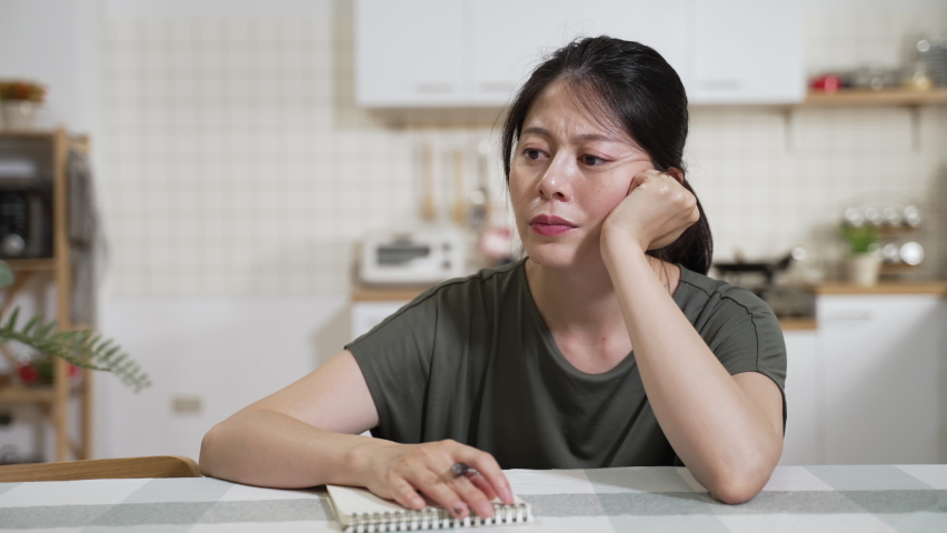 Front portrait of a stressed asian wife tapping hand on the notebook while planning and worrying about family financial problems at dining table in the apartment | Shutterstock HD Video #1093857355
