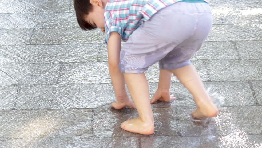 water falls on sidewalk brick wall road,child kid is playing jumping having fun in sunny summer day.barefoot boy runs touching the road with palm,splashing.cascate waterfall in city for heat days,cool