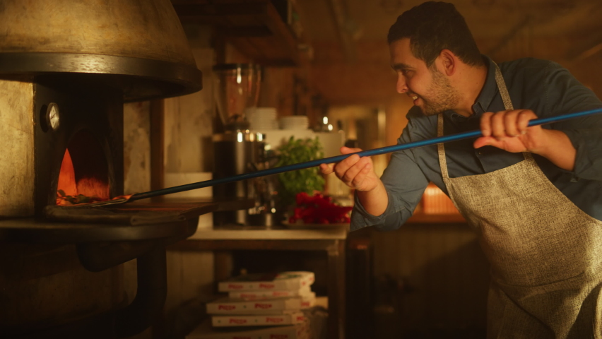 In Restaurant Professional Chef Uses Pizza Peel for Transferring Pizza into Wood Fire Stone Oven. Traditional Italian Cooking Family Recipe. Authentic Pizzeria, Delicious Organic Food. Cinematic Shot
