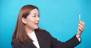 Young happy Asian businesswoman using smartphone to selfie smiling waving in a black suit on blue background
