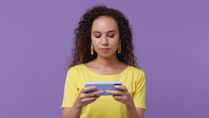 Young gambling woman of African American ethnicity 20s wear yellow t-shirt using play racing on mobile cell phone hold gadget smartphone for pc video games isolated on plain pastel purple background