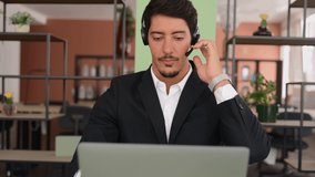 Front view at smiling latin male employee wearing headset and smart casual wear using laptop for connection with colleagues or customers, talking online sitting near windows in the office