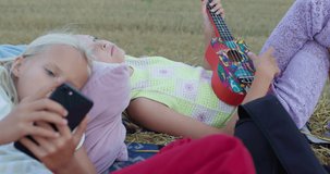 charming girl playing the ukulele on mown rye in field. Landscape of straw bales against setting sun on background. Happy sisters watch video on smartphone outdoors. Childhood. Country life