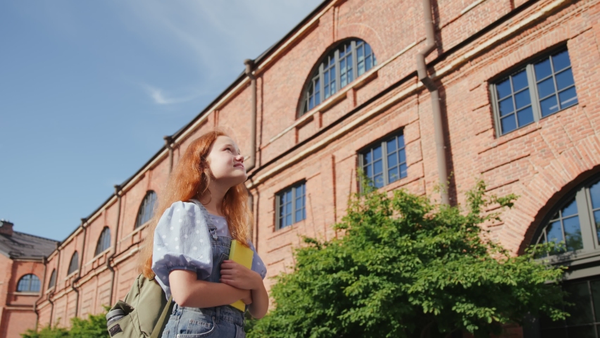 Low angle view of smiling teenage girl with book and backpack standing outside school building, looking around and smiling. Education, back to school concept
