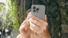 Close-up of woman shooting video on modern phone while standing under waterfall on sunny day. Girl hides her face behind her phone while standing in jungle under streams of water of mountain river