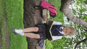 Vertical video. Schoolgirl doing homework while sitting on a tree branch in a park, reading interesting book. Back to school, education concept