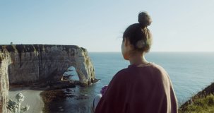 A beautiful young woman sits on a hilltop, admiring a beautiful view of the picturesque sheer cliffs with a natural arch, located on the banks of English Channel. Etretat, France. 4k video, red komodo