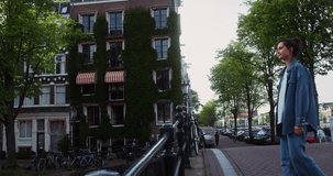 A young woman admires the views of a beautiful European city, standing on a bridge over a canal. Lots of bicycles on shore near house overgrown with ivy. Amsterdam, Netherlands, 4k video, red komodo
