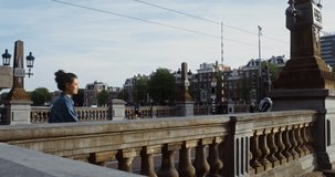 A beautiful young woman leans on the parapet of the bridge, enjoying the panorama of the beautiful European city on the water with a smile. Amsterdam, Netherlands. Video in 4k, red komodo