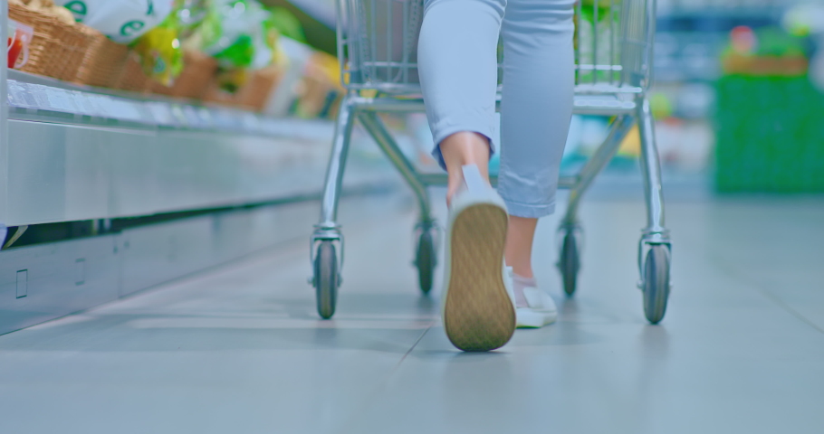 Women's feet in sneakers walking around the supermarket. Woman drives a grocery cart through a supermarket, rear view. 4k, ProRes Royalty-Free Stock Footage #1093968441