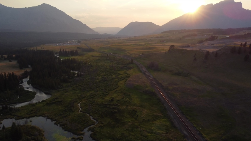 High aerial shot over the winding Crowsnest River with distant Canadian Rocky Mountains and train tracks in Alberta Canada at sunset.
