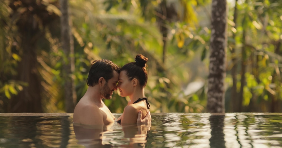 Panoramic photo of young adult couple in love swimming together in open-air infinity pool enjoying tropical vacation on villa looking to each other against rainforest on sunset. High quality 4k Royalty-Free Stock Footage #1093969783