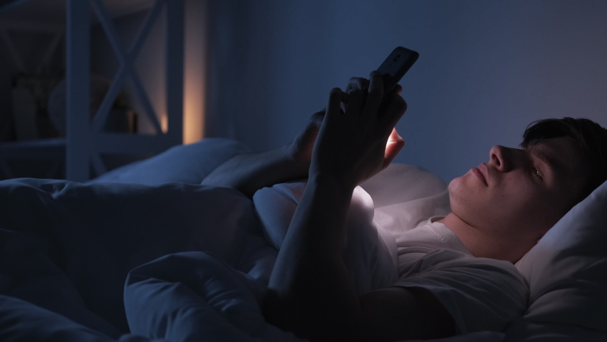 Night chat. Internet fatigue. Social media addiction. Tired exhausted bored man falling asleep using phone in bed late in dark bedroom.