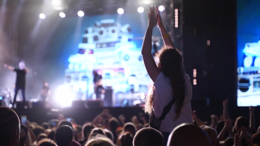Woman sits on boyfriend shoulders waving hands at music concert on open air fest. Crowd applause, jump, wave hands and dance at favourite band performance on stage. Bright illumination. Royalty-Free Stock Footage #1093996417