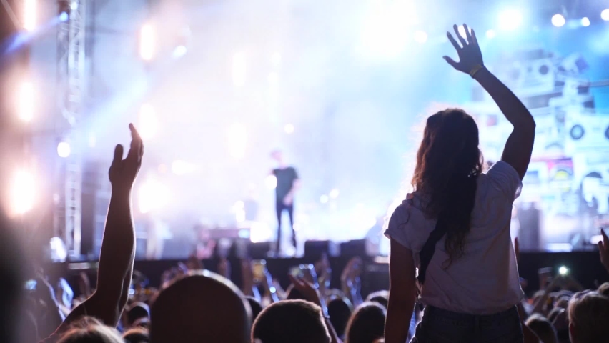 Woman sits on boyfriend shoulders waving hands at music concert on open air fest. Crowd applause, jump, wave hands and dance at favourite band performance on stage. Bright illumination.