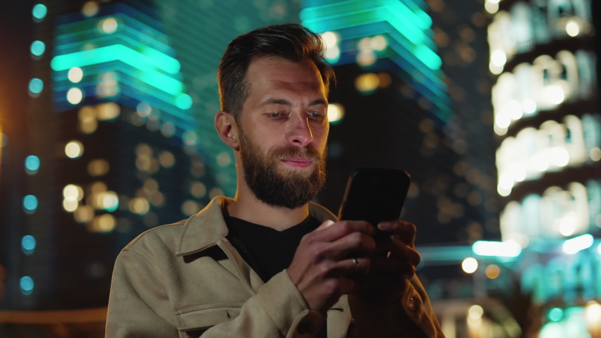 One Man with Phone in Hand at Dark Urban Street Sharing News. Person Walks in Neon Evening and Uses Smartphone Near Futuristic Illuminated Buildings. Electronic Information for Big Life of Traveler