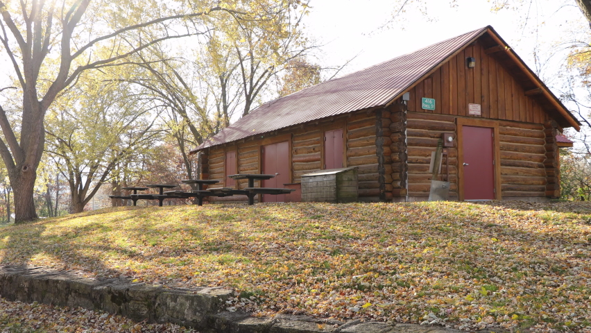 Autumn view of log built park shelter on a beautiful autumn day. Sun creating shadows across the leaf-covered grass. Picnic tables along the side. Red metal roof. Tranquil nature scene.  