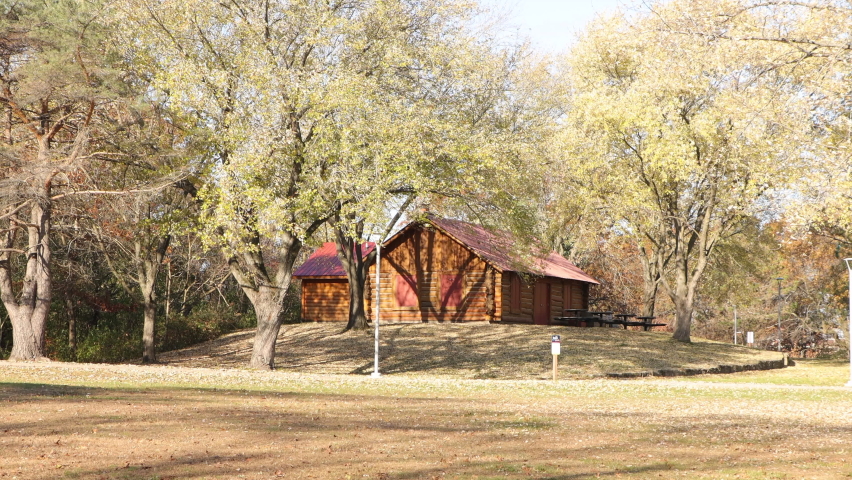 Beautiful autumn day view of a log built park shelter. Leaves all over the grass and dirt. Trees with leaves and bare branches. Shadows cast from the bright sun. Picnic tables by shelter.