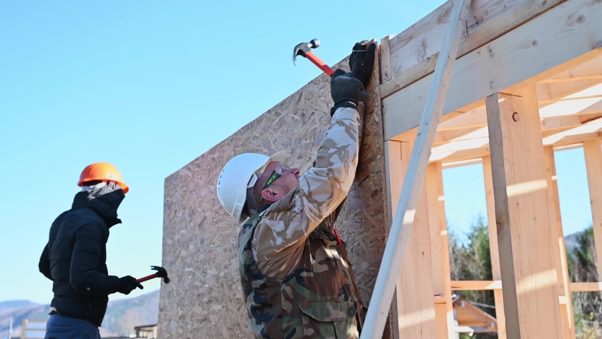 Carpenters hammering nail into OSB panel on the wall of future cottage. Men workers building wooden frame house. Carpentry and construction concept. Royalty-Free Stock Footage #1094003367