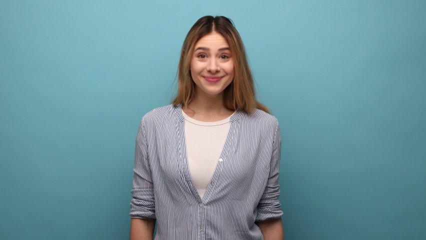 Positive optimistic woman looking at camera and nodding yes, showing approving gesture, body language concept, wearing striped shirt. Indoor studio shot isolated on blue background.
