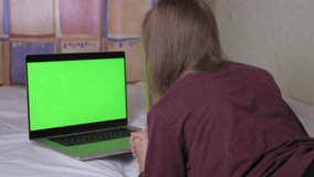 A cute teenage girl is lying on the bed, in front of her is a laptop with a green screen. A teenager is alone in her room looking at a laptop screen.