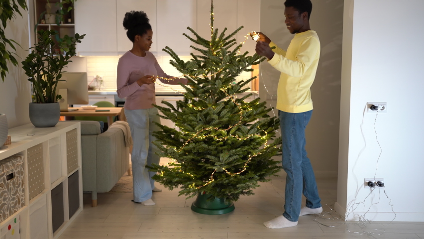 Young african family couple create minimalist Christmas tree by skipping ornaments to celebrate winter holidays in minimal way, black man and woman decorate xmas tree together with only twinkle lights