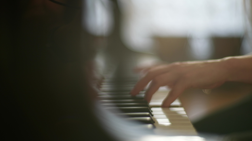 Close-up tracking shot of unrecognizable talented young woman pianist playing gentle music on beautiful grand piano sitting on background of window, slow motion, selective focus in rays of sun.