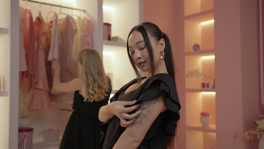 Two women friends choosing new outfit standing in front of the mirror in dressing room. Women in black dresses trying on various dresses in wardrobe. Royalty-Free Stock Footage #1094029955