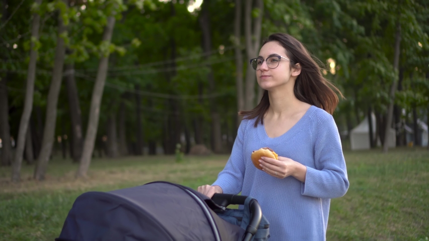 A young woman in glasses walks with a stroller in the park and eats a burger. Mom walks with a child in nature close-up.