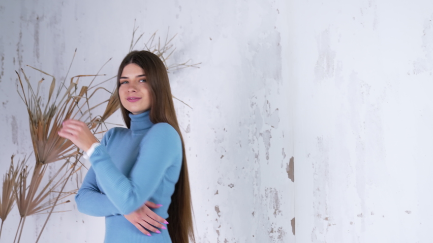 Beautiful long-haired woman in warm light blue dress indoors. Fashionable lady in tight dress and white boots on tractor soles. White backdrop.
