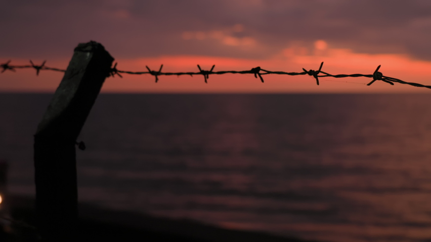 Dark silhouette of barbed steel wire on pole making a restriction fence against amazing dusk on ocean and a passing motorcycle. Obstacle preventing from dream coming true metaphor. Symbol of banned.