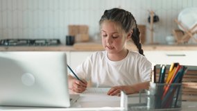 Teenage schoolgirl doing homework at table with a laptop. Distance education home. Child uses gadgets to study. Education and entertainment at home. School at home kitchen table. Modern technologies.