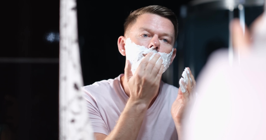 Closeup of reflection in mirror of confident handsome young man and applying shaving cream