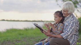Happy smiling old grandmother reding funny book fairy tale, embracing little joyful granddaughter sitting on lake beach outdoors. Child thinking and imagination development, calm family rest at nature - Powered by Shutterstock - Get 15% off with code: PIKWIZARD15
