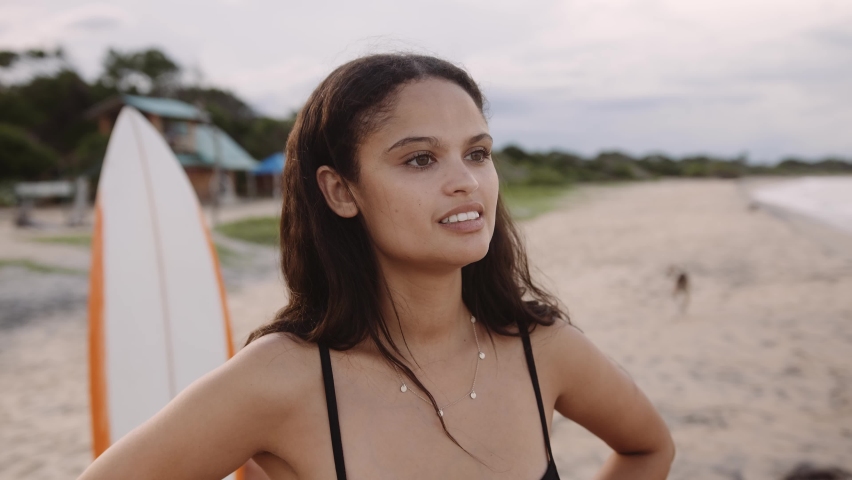 Happy young biracial woman picking up surfboard on beach by sea | Shutterstock HD Video #1094063023