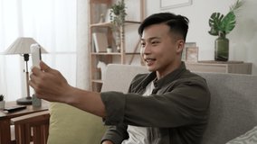 smiling asian man staying at home in casual wear is talking happily while having a virtual chat with friends on the smartphone in the living room.