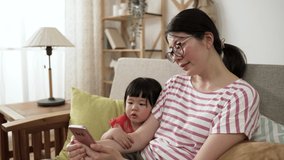 cute asian toddler baby girl snuggling with mother on sofa in living room while watching educational video for kid on the internet with phone together at home.
