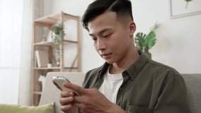 closeup shot of a happy asian guy having hearty laugh while enjoying funny social media feeds on smartphone in a modern living room at home with daylight.