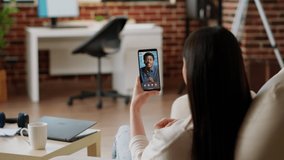 Employee working remotely from home while using talking with coworker on virtual online videoconference call. Young adult woman discussing with work colleague on digital videocall using phone.
