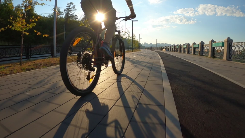 Aerial low-angle back view from FPV drone to cyclist riding on mountain bike on bike trail at city embankment in early morning sunrise on background of bright sunlight. Tracking shot in slow motion.