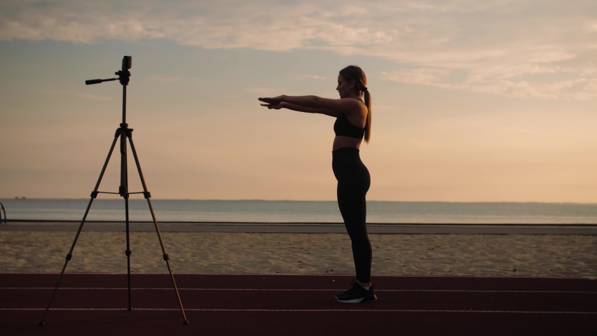 Young Woman in sportwear doing fitness online course.Girl doing back flip, acrobatic activity at sunrise. Woman teaching fitness online on the beach. High quality 4k footage