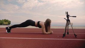 Fitness girl standing in the plank.Woman teaching online using smartphone on the playground.Training outdoors on the beach. High quality 4k footage