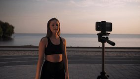 Fitness blogger girl doing fitness exercises recording on the phone.Woman recording online course about stretching against the backdrop of the sea and the beach. High quality 4k footage