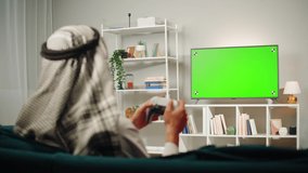 Middle eastern man playing video game, tv with chroma key. Television with green screen, play gaming on modern console. Wearing traditional Islamic male clothes. 