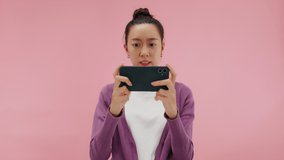 Attractive young asian woman holding smartphone and enjoy playing mobile game application. Beautiful girl focused looking at gadget cell phone felling exited and stress. isolated on pink background