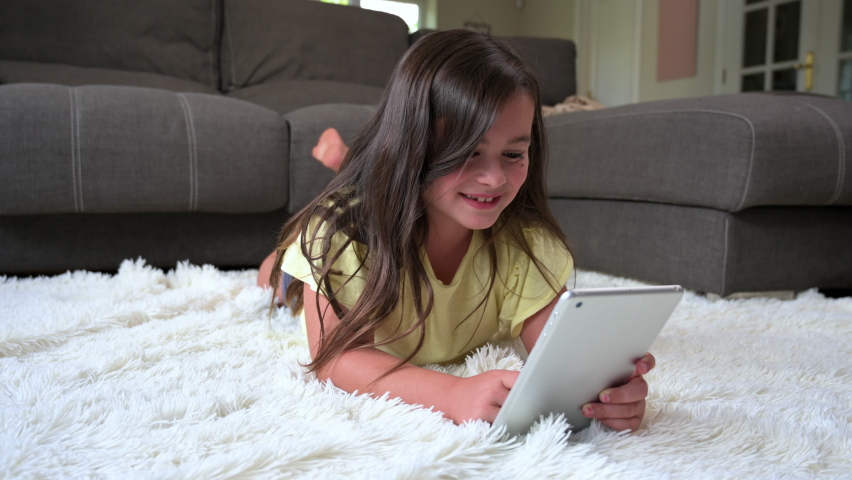 Cute little girl watch cartoons on digital tablet. Kid lies on floor laughing using electronic device. Indoor leisure for children. High quality 4k footage.
