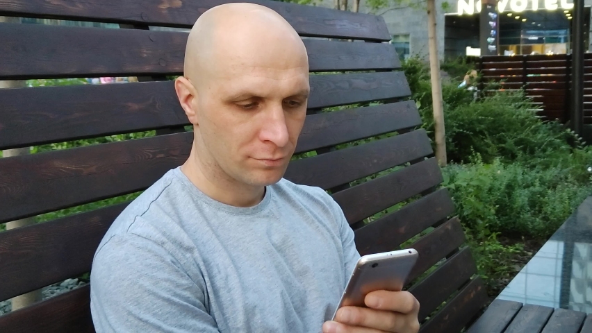 White Caucasian man reads news on his smartphone. Handsome bald guy look at mobile phone screen. Using  cell phone for work and leisure. Installing application. Modern technologies. Close-up, portrait