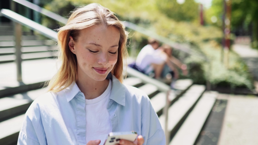 Girl university student social media user holding cell phone using mobile applications tech on smartphone looking at cellphone, chatting, texting, checking educational apps standing outside campus. Royalty-Free Stock Footage #1094083531