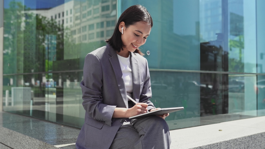 Happy elegant young Asian business woman executive, professional manager wearing suit holding using digital tablet modern fintech smart device working online sitting in big city outdoors. Royalty-Free Stock Footage #1094083541