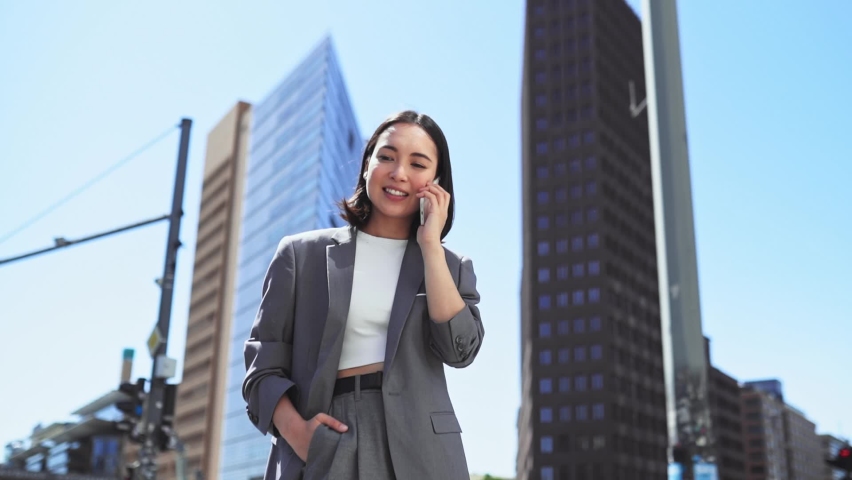 Young happy Asian successful businesswoman wearing suit standing on big city street talking on mobile phone. Smiling woman making business call on cellular on sky urban buildings, slow motion. Royalty-Free Stock Footage #1094083551